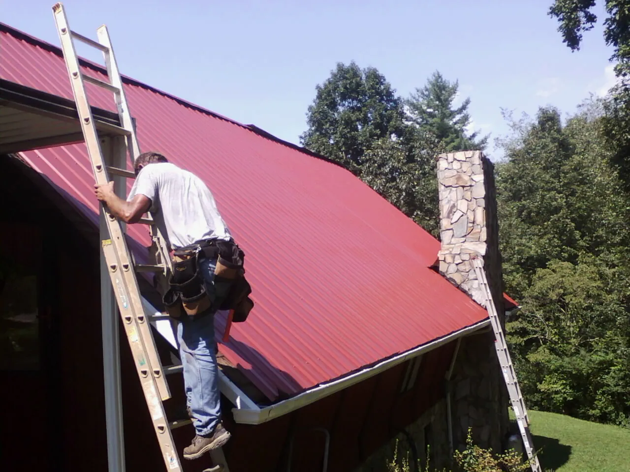 Roofer on a ladder beside a red metal roof and stone chimney — Soffit & Fascia Repair by Roofing Brothers in Palmdale